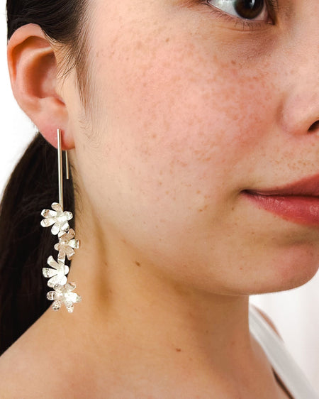 Die Ohrringe „Jacintos“ verbinden schlichte Eleganz mit handwerklicher Präzision. Mehrere kleine, von Hand geformte Blüten hängen aneinander und erzeugen bei jeder Bewegung ein feines Spiel aus Glanz und Struktur.