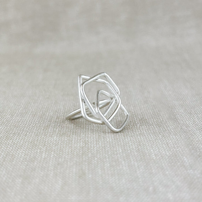 Ein moderner Ring mit klarer, architektonischer Formensprache. Handgefertigt aus feinem 100% recyceltem Sterlingsilber 925, besticht dieses Schmuckstück durch seine leichte, offene Struktur aus ineinander verschlungenen Quadraten. Die matte Oberfläche verleiht dem Ring eine sanfte, elegante Ausstrahlung – zeitlos und doch auffällig.